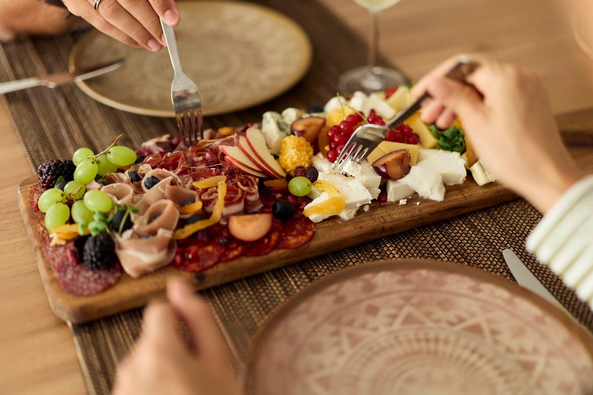 Hands lifting cheese, cured meats and fresh fruit from a shared charcuterie board
